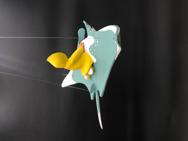 Vu du derrière de la céramique du personnage en ciré jaune et lunnettes de coeur rouges sur une manta. La pièce a des fils pour l'accrocher au plafond.