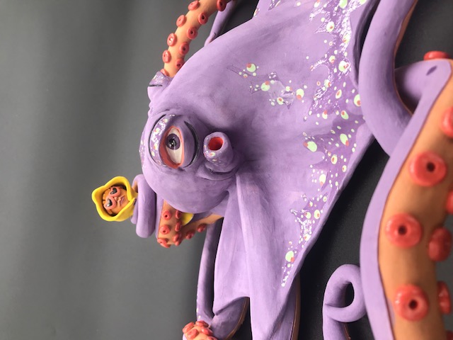 Pièce en céramique d'un poulpe mauve qui tient dans un de ses tentacules le personnage au ciré jaune et lunnettes de coeur.