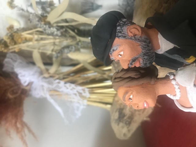 Détail de la pièce en céramique qui représente un couple de mariés, homme et femme. La sculpture a des fleurs sèches derrière.