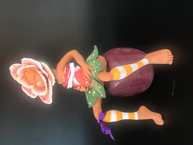 Céramique de fleur, au milieu une tête de femme. Elle est assise sur un pouf et touche son ventre. Sa jupe est faite de feuilles.