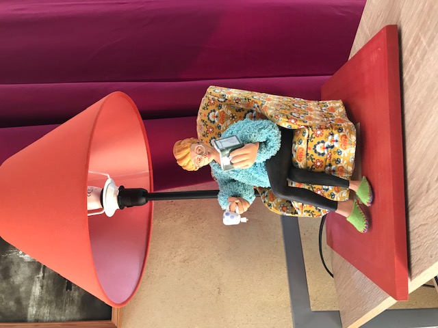 Lampe rouge. Sur la base, il y a une pièce de céramique qui représente une dame à lunettes et pantoufles assise sur un fauteuil à fleurs. Elle lit un livre et a une tasse de thé à l'autre main.