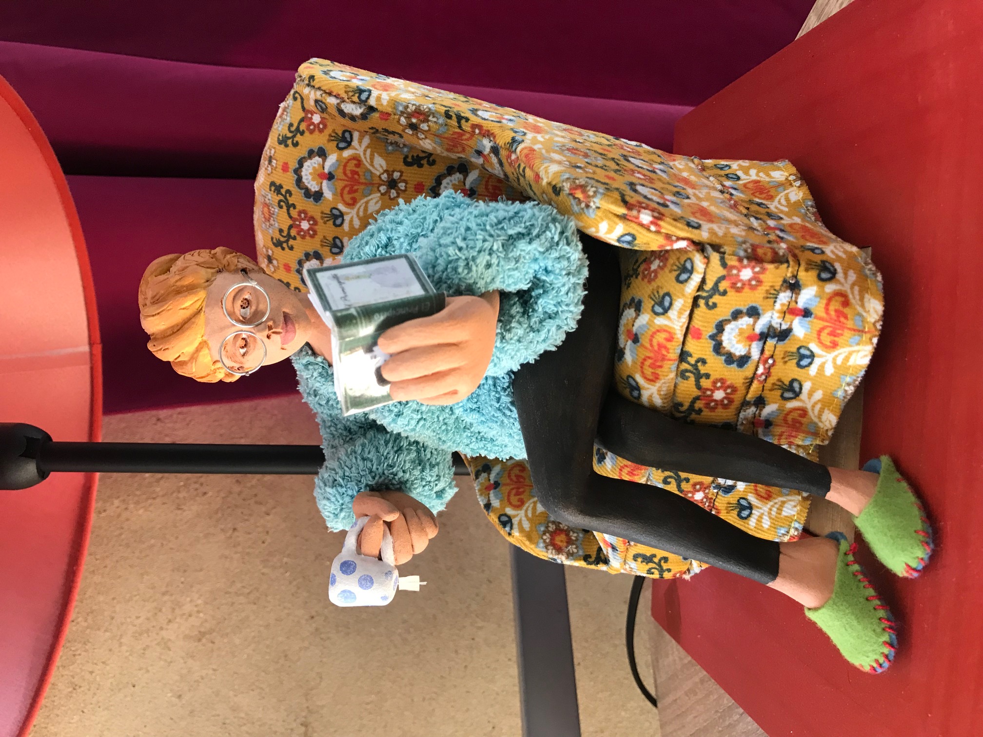 Base de la lampe rouge, il y a une pièce de céramique qui représente une dame à lunettes et pantoufles assise sur un fauteuil à fleurs. Elle lit un livre et a une tasse de thé à l'autre main.