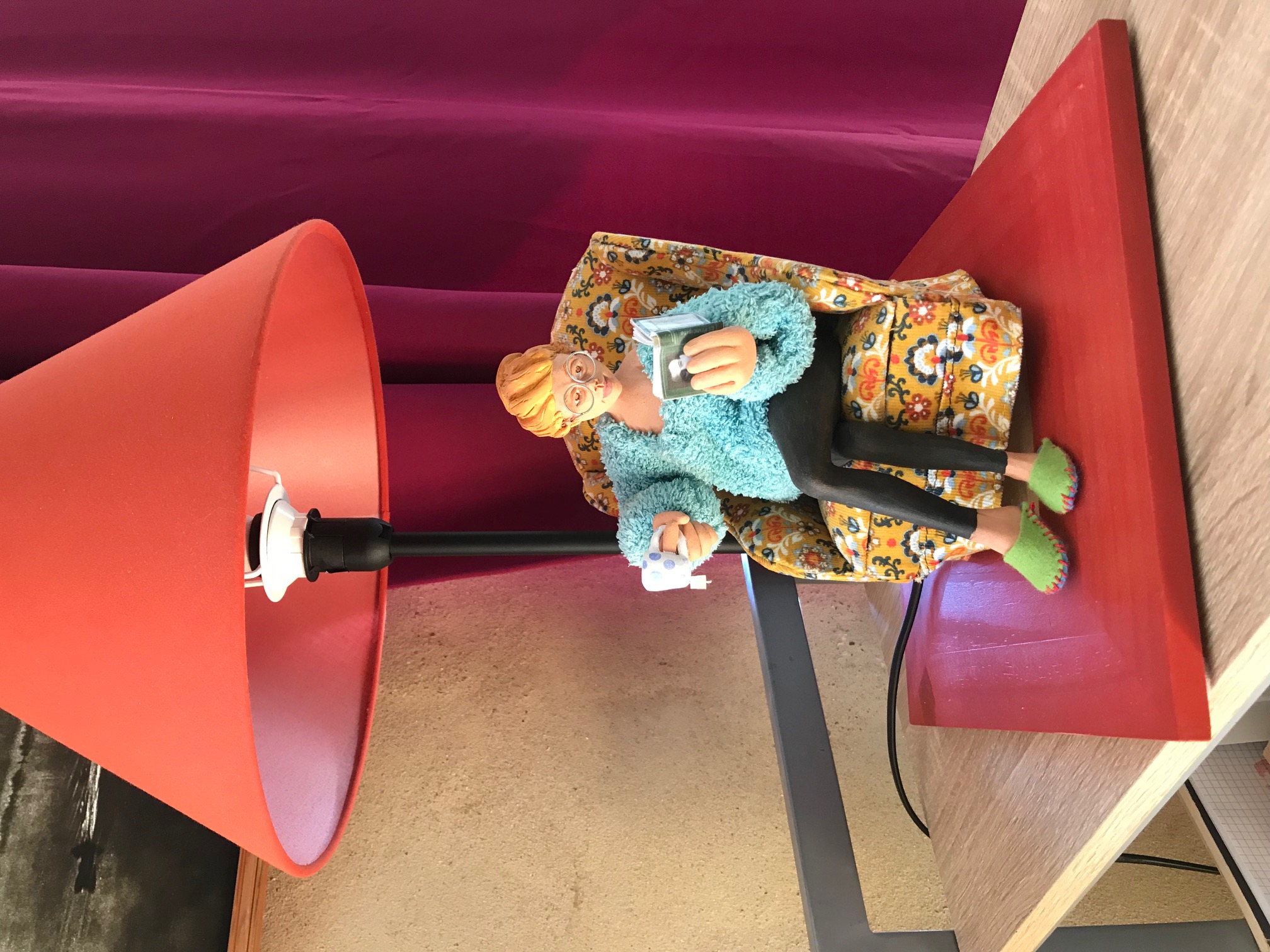 Lampe rouge. Sur la base, il y a une pièce de céramique qui représente une dame à lunettes et pantoufles assise sur un fauteuil à fleurs. Elle lit un livre et a une tasse de thé à l'autre main.