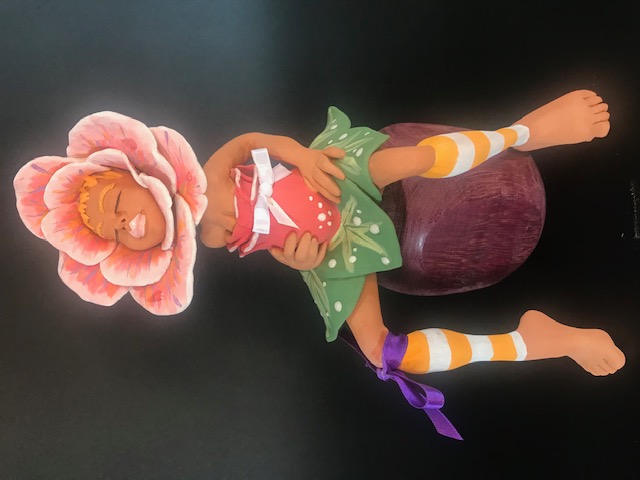 Céramique de fleur, au milieu une tête de femme. Elle est assise sur un pouf et touche son ventre. Sa jupe est faite de feuilles.