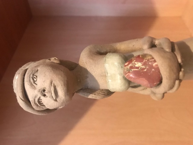 Détail de la sculpture en céramique d'une femme mince à la robe longue. Elle a un ruban aux cheveux et regarde vers le haut. El porte dans ses mains un bol avec un minéral