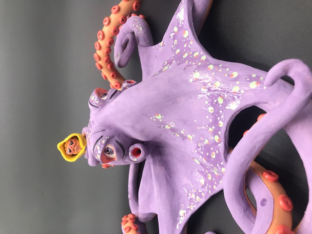 Pièce en céramique d'un poulpe mauve qui tient dans un de ses tentacules le personnage au ciré jaune et lunnettes de coeur.