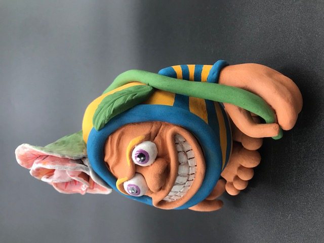 Pièce en céramique Tête d'Oeuf. Un personnage qui montre toutes ses dents en riant, il porte une fleur sur sa tête, qui occupe presque tout son corps. Il tient la tige de la fleur avec sa main gauche.