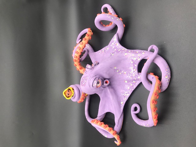 Pièce en céramique d'un poulpe mauve qui tient dans un de ses tentacules le personnage au ciré jaune et lunnettes de coeur.