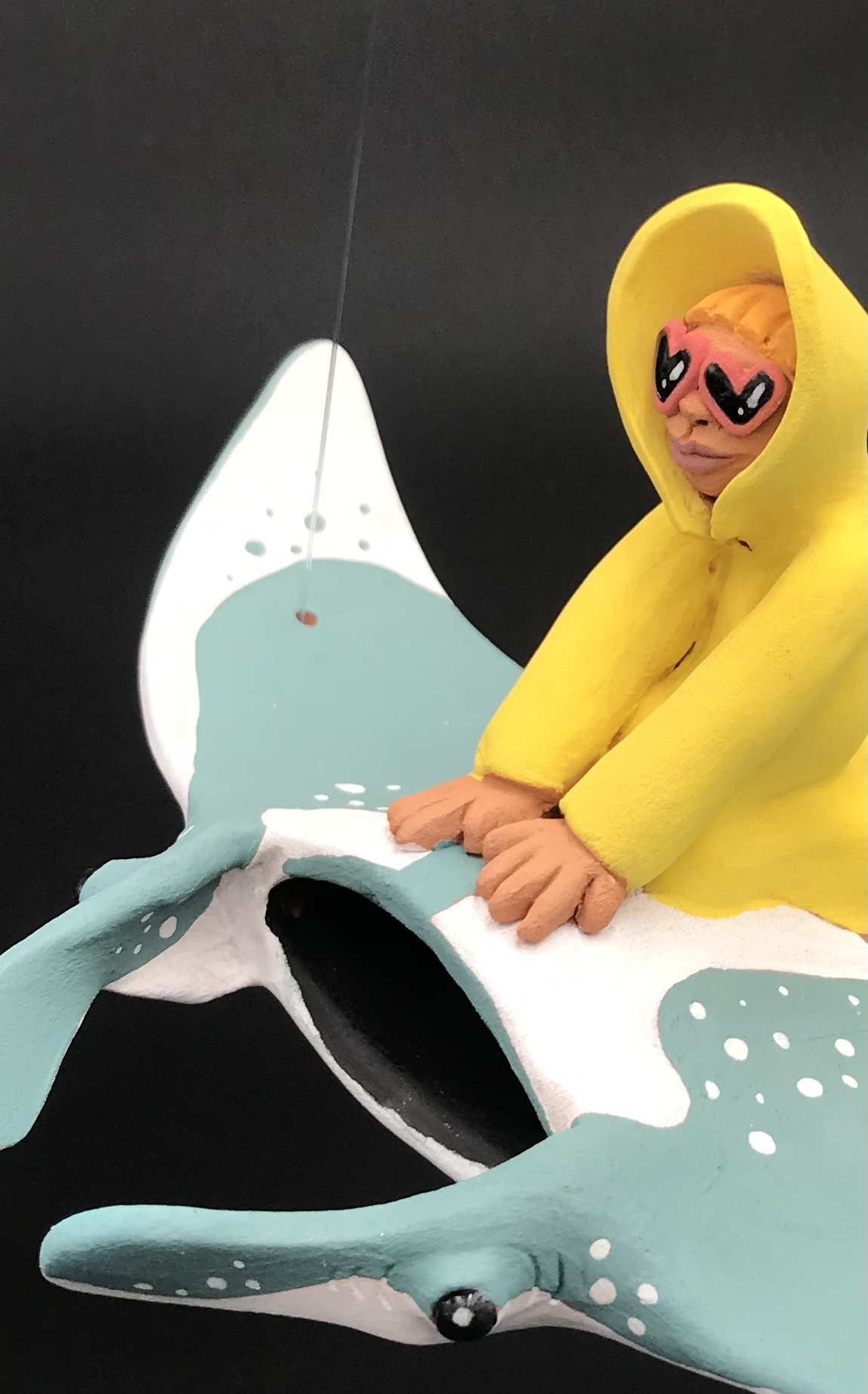 Détail de la céramique du personnage en ciré jaune et lunnettes de coeur rouges sur une manta. La pièce a des fils pour l'accrocher au plafond.