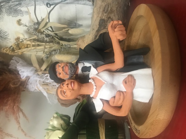 Pièce en céramique qui représente l'image de tête à ceinture d'un couple de mariés, homme et femme. L'homme prend la femme par la taille, ils serrent ses mains. Au fond il y a des fleurs sèches.