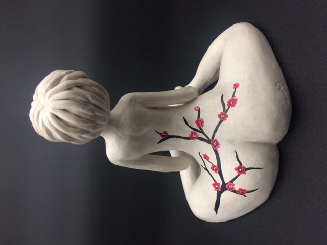 Sculpture en céramique d'une femme aux cheveux courts nue et assise. Elle a un tatuage d'une branche de cerisier qui va de sa jambe gauche au dos.