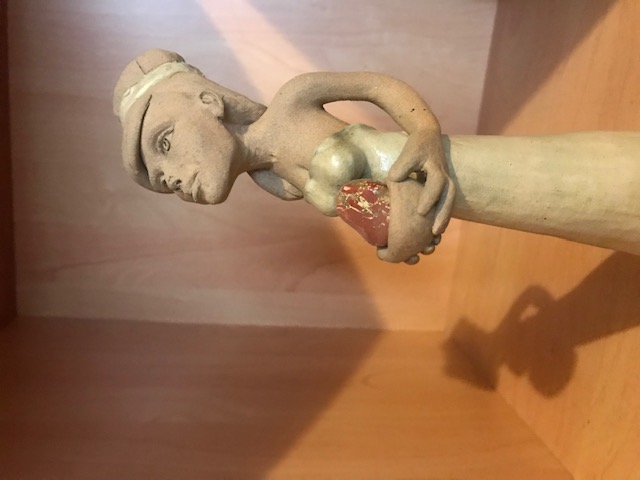Petite sculpture en céramique d'une femme mince à la robe longue. Elle a un ruban aux cheveux et regarde vers le haut. El porte dans ses mains un bol avec un minéral