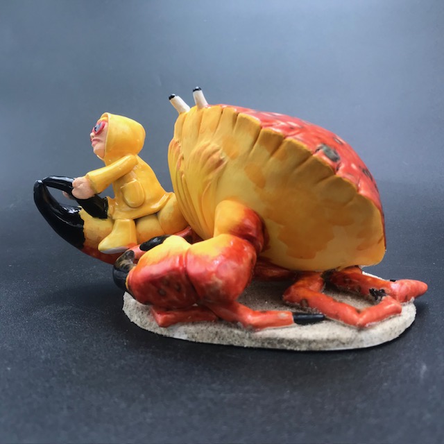 Sculpture en céramique d'une petite fille en ciré jaune à califourchon sur la pince d'un crabe
