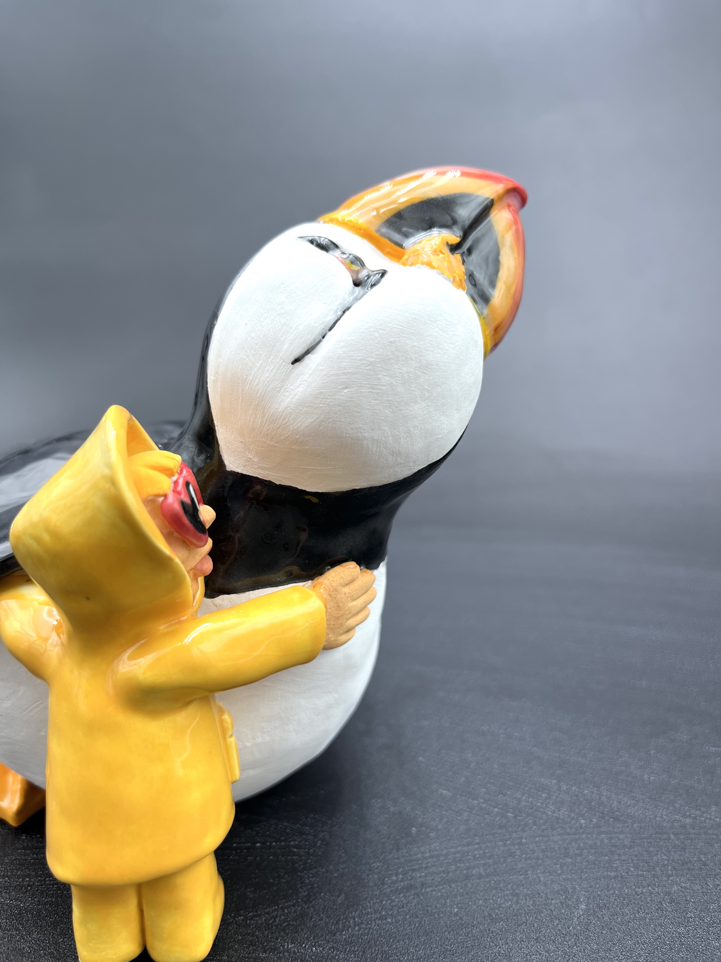 Sculpture en céramique d'un petit personnage en ciré faisant un calin à un macareux